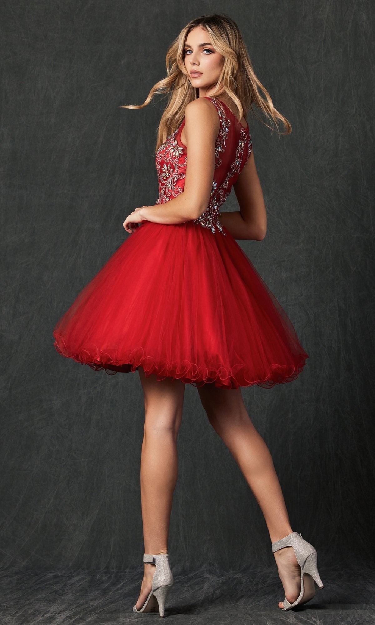A woman poses in high heels against a dark background, wearing the Short Hoco Dress: Juliet 749—a sleeveless red homecoming dress with an embellished bodice and A-line tulle skirt.